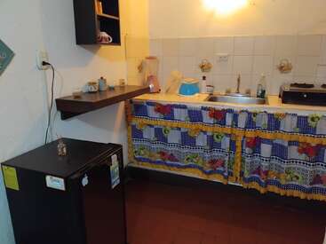 Dieses Bild zeigt eine kleine, gemütliche Küche mit einem Mini-Kühlschrank, einem bunten Vorhang unter der Spüle, Holzregalen, gefliesten Wänden und verschiedenen Küchenutensilien.