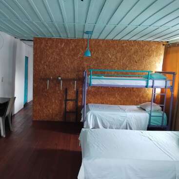 Dieses Bild zeigt ein einfaches Herbergszimmer mit weißem Bettzeug, Etagenbetten, Holzböden, einer hölzernen Trennwand, einer blauen Decke und minimalem Mobiliar, darunter Stühle und Regale.