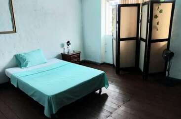 Es ist ein einfaches Schlafzimmer mit mintgrüner Bettwäsche, Holzboden, einem Beistelltisch mit einer Lampe, einem Fenster und einem klappbaren Sichtschutz neben einem Ventilator.