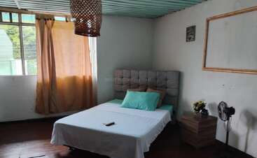 Ein gemütliches Schlafzimmer mit einem ordentlich gemachten Bett, einem hellen, tealfarbenen Kissen, einem Holzboden, einem Nachttisch mit Blumen, einem Fenster mit Vorhängen, einem Ventilator und Retro-Deko-Akzenten. "Casa Retro"-Logo.