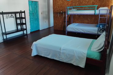 Esse quarto tem uma cama de solteiro, beliches, prateleiras abertas com cabides, piso de madeira, porta turquesa e uma decoração minimalista, criando um ambiente simples e organizado.