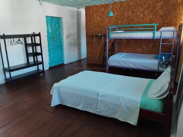 This room features a single bed, bunk beds, a teal door, open shelving with hangers, wooden floors, and a pendant light. Simple, minimalist style.