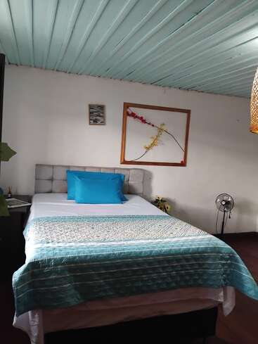 This cozy bedroom features a neatly made bed with teal bedding, light blue ceiling, simple wall art, small bedside tables, a fan, and wooden flooring.