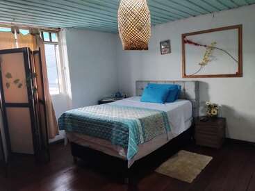 A cozy bedroom with a double bed, blue bedding, wooden floor, wicker ceiling lamp, window with curtains, side table, and decorative art on the wall.