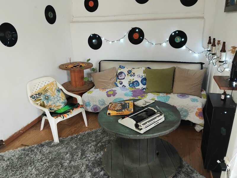 This cozy room features a floral couch, vinyl records on white walls, string lights, books, a typewriter on a spool table, and a white plastic chair.