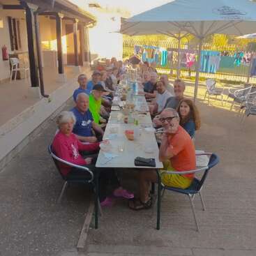 A large group of people are gathered around a long outdoor table, enjoying a meal together. There are adults and seniors, with a relaxed, cheerful atmosphere.