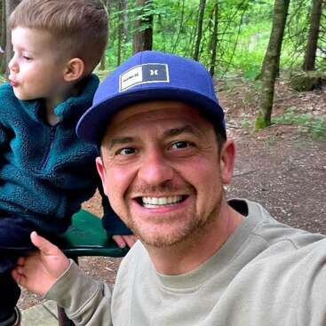 Ein lächelnder Mann mit einer blauen Mütze macht ein Selfie im Wald und hält ein Kind, das wegschaut. Bäume und Grünflächen umgeben sie im Freien.