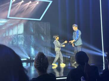 A magician on stage interacts with a young boy under dramatic lighting. Audience members watch from the foreground, enjoying the live performance in a theater setting.