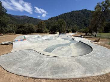 This image shows a large, concrete skate park surrounded by nature, mountains, and trees under a blue sky with scattered clouds. People are skating and relaxing nearby.