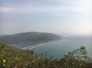 The image depicts a serene coastal scene with a misty sky, featuring a body of water, a sandy beach, and lush greenery in the foreground.