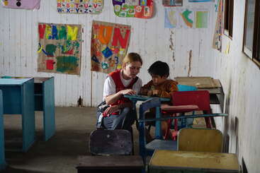 Une femme aide un jeune garçon à faire ses devoirs dans une salle de classe remplie de dessins colorés, de bureaux dépareillés et d'affiches éducatives sur les murs.