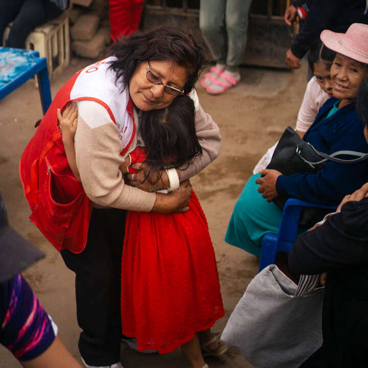 A imagem mostra uma mulher com um avental vermelho abraçando uma menina com um vestido vermelho, cercada por um grupo de pessoas em um ambiente externo.