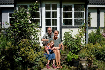 Una familia de tres miembros posa sonriente frente a una casa rodeada de exuberante vegetación y flores, disfrutando juntos de un día soleado e irradiando calidez y felicidad.