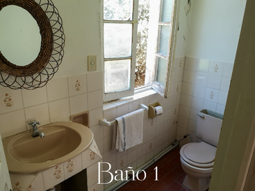 Esta imagem mostra um banheiro pequeno com uma pia bege, espelho redondo, paredes azulejadas, vaso sanitário, porta-toalhas, janela aberta e o texto "Baño 1" exibido.