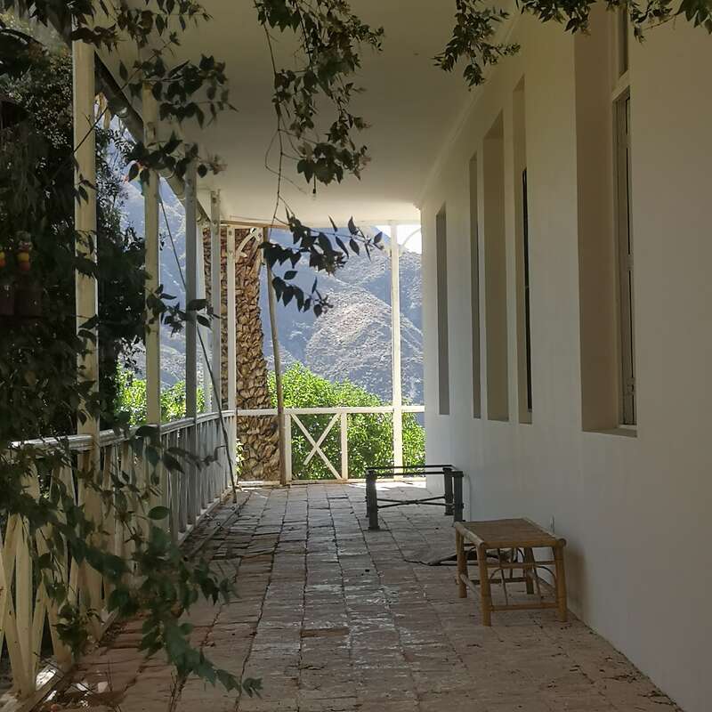 Uma varanda tranquila e iluminada pelo sol, com piso de ladrilhos, grades brancas, um banco simples, trepadeiras frondosas no alto e uma bela vista da montanha ao fundo. Uma atmosfera muito serena.
