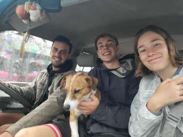 Três pessoas sorridentes sentam-se juntas dentro de um carro em um dia chuvoso, acompanhadas por um cachorro marrom e branco. A atmosfera é amigável, calorosa e aventureira.