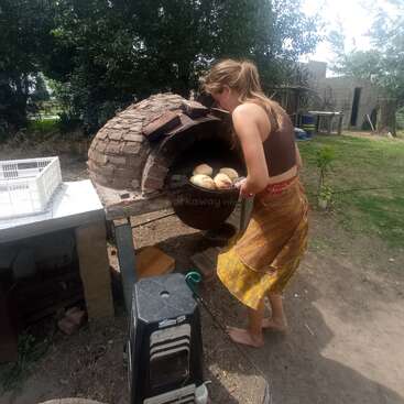 Une femme fait cuire du pain dans un four extérieur en briques. Elle est pieds nus, vêtue d'un haut marron et d'une jupe jaune, entourée d'un jardin rustique et verdoyant.