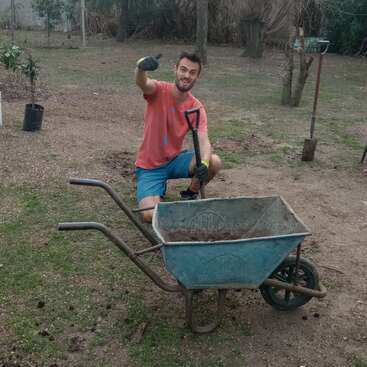 Un homme est agenouillé sur le sol, souriant et levant le pouce alors qu'il travaille dans un jardin avec une brouette et une pelle par une journée nuageuse.