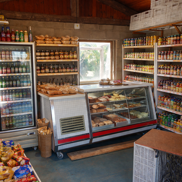 Ein kleiner, uriger Laden mit Regalen voller Konserven, einem Kühlschrank mit Getränken, einer Auslage mit verschiedenen Brotsorten und Backwaren sowie Snacks und einer Holzdecke.