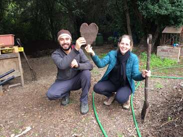 Duas pessoas sorriem e posam em um jardim, segurando um objeto em forma de coração e uma pá, cercadas por ferramentas, vegetação e uma atmosfera alegre e divertida.