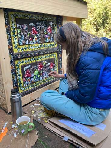 Uma mulher senta-se de pernas cruzadas no chão, pintando desenhos intrincados e coloridos de casas e cogumelos em uma estrutura de madeira. Perto dela há uma caneca e uma garrafa térmica.