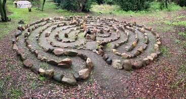 Esta imagem mostra um labirinto em espiral feito de pedras dispostas no chão em um ambiente natural ao ar livre, cercado por árvores e vegetação, evocando uma meditação pacífica.
