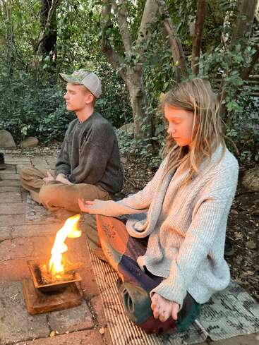 Duas pessoas sentam-se com as pernas cruzadas ao ar livre em um ambiente sereno de floresta, meditando em silêncio ao redor de uma pequena fogueira em um tapete, cercadas por vegetação e ambiente tranquilo.