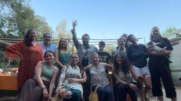 Um grupo de quatorze pessoas, algumas sentadas e outras em pé, sorriem ao ar livre. Uma pessoa levanta a mão com entusiasmo. Duas pessoas seguram animais de estimação. Árvores e mesas os cercam.