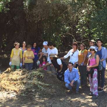 Um grupo de pessoas, incluindo adultos e crianças, se reúne ao ar livre em torno de uma pilha de compostagem. Elas parecem alegres e envolvidas em uma atividade na natureza ou de jardinagem.