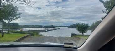 Viewed from inside a car, the scene overlooks a serene river with a pier, surrounded by trees, grassy banks, and under a partly cloudy sky. Peaceful atmosphere.