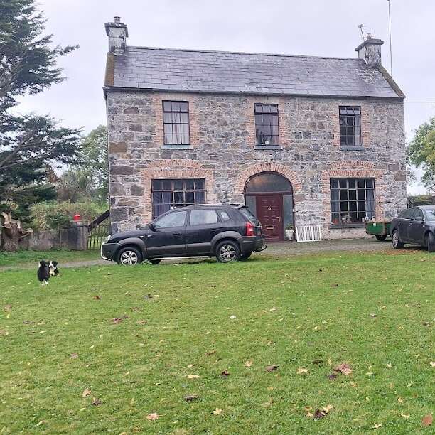A two-story stone house with large windows stands on a grassy lawn. Two cars are parked in front, and a black-and-white dog is nearby.