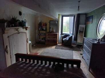 A cozy, rustic bedroom with wooden floors, soft rugs, a bed, dresser, armchair by the window, plants, bookshelves, and mirrors, bathed in natural light.