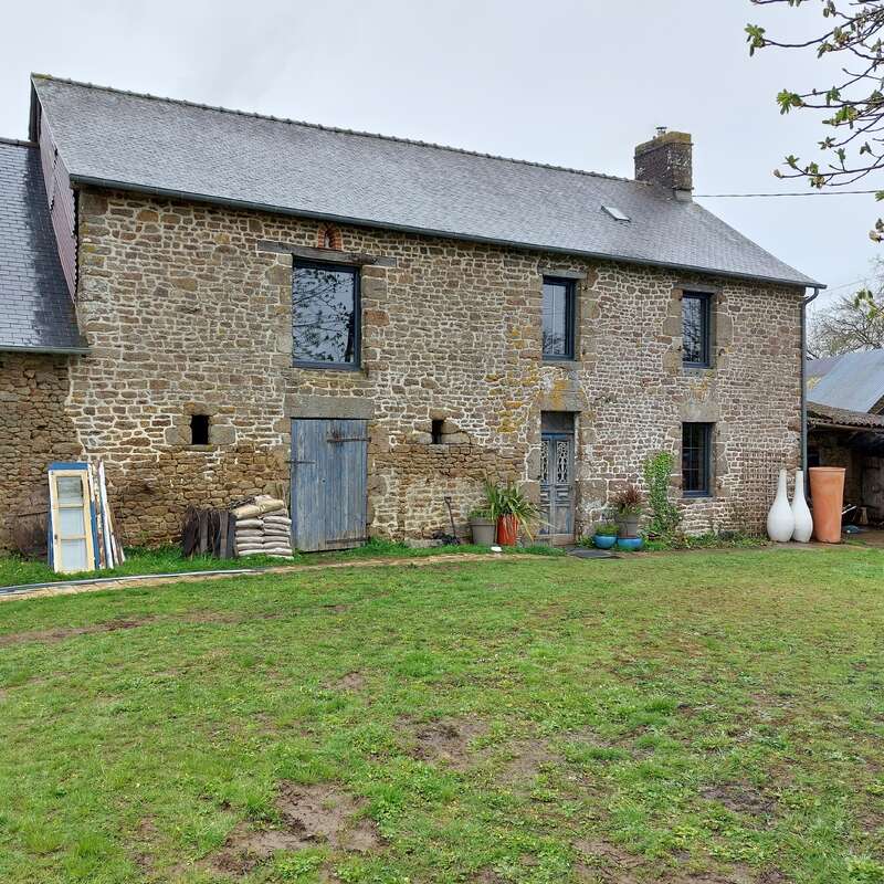 A rustic stone house with blue doors and windows stands on a grassy yard. Potted plants, old windows, and large vases decorate the front.