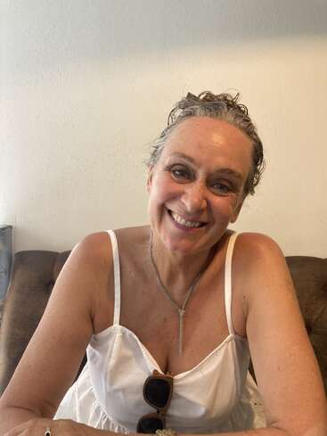 A smiling woman with curly hair sits on a brown couch, wearing a white dress and silver necklace. She looks cheerful and relaxed, sunglasses in hand.