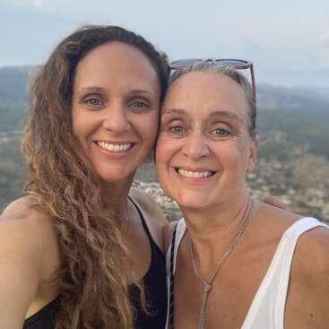 Two women smile warmly while taking a selfie outdoors. They appear close, possibly family. A scenic landscape with hills and sky provides the background, adding tranquility and depth.