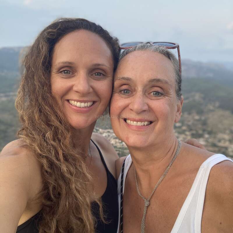 Two women smile warmly while taking a selfie outdoors. They appear close, possibly family. A scenic landscape with hills and sky provides the background, adding tranquility and depth.