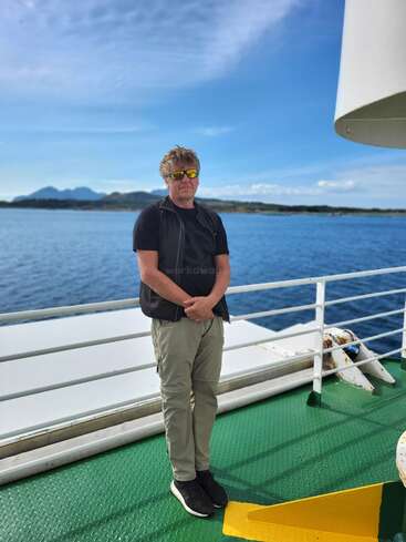 A man wearing sunglasses stands on a ferry’s deck, with blue sea and distant mountains behind him under a bright, mostly clear sky, enjoying the view.