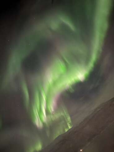 Swirling green aurora borealis dances across a dark night sky above a barren landscape, with faint stars visible through the luminous, ethereal light curtains. Mesmerizing natural display.