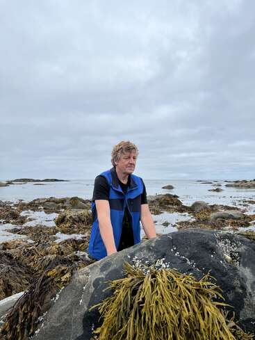 A man in a blue vest stands among seaweed-covered rocks on a cloudy day by the seaside, looking thoughtful. The ocean and sky appear calm.
