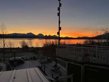A beautiful sunrise or sunset over a calm lake with mountains in the background, bare trees, frosty ground, a fence, and outdoor furniture in the foreground.