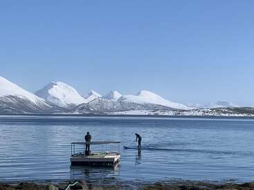 A person stands on a floating platform while another paddleboards on a calm lake, surrounded by snow-covered mountains under a clear blue sky. Peaceful, scenic landscape.