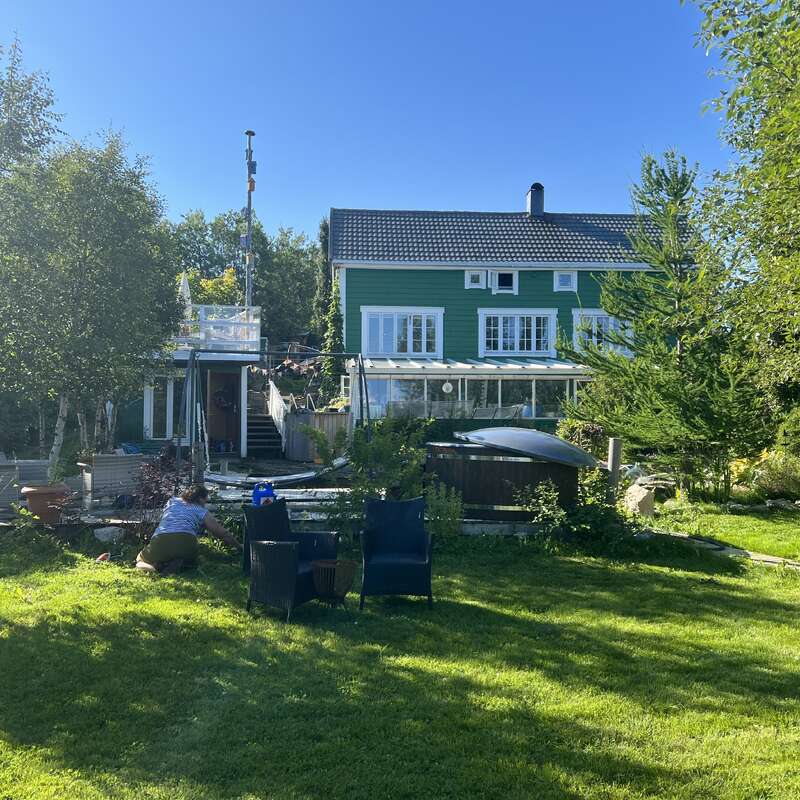 A green house with a glass veranda, surrounded by lush trees and garden. A person tends plants, with outdoor furniture on the sunlit lawn. Clear blue sky.