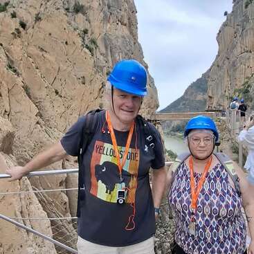 A man and woman wearing blue helmets, lanyards, and backpacks stand on a scenic bridge with rocky cliffs and a river gorge in the background.