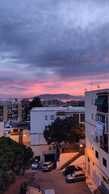 Esta imagem mostra uma cidade ao pôr do sol, com belas nuvens rosa e roxas acima. Prédios brancos e amarelos, carros estacionados e árvores preenchem a paisagem urbana.