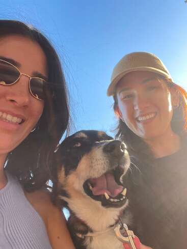 Duas mulheres sorridentes, uma usando óculos escuros e a outra um boné bronzeado, posam ao ar livre com um cachorro feliz e de boca aberta em um dia ensolarado.