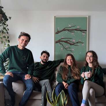 Four people sit on a cozy couch in a bright, modern living room with green plants, art of tree branches, and all wearing green sweaters, smiling warmly.