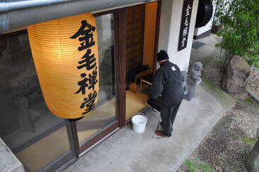 A man stands outside a traditional Japanese building, holding a bucket. A large paper lantern with Japanese characters hangs, and a stone pathway and plants are visible.