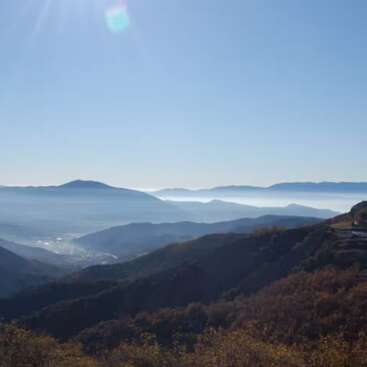 This serene image shows layers of distant mountains under a clear blue sky, lightly veiled with morning mist, bathed in gentle sunlight. Nature feels peaceful and vast.