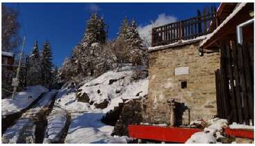 A snowy mountain scene features a rustic stone building, wooden fence, and road with tire tracks, surrounded by snow-covered trees and a bright blue sky above.
