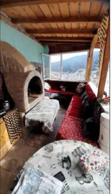 This cozy indoor patio features a brick oven, two tables with printed tablecloths, red cushioned bench seating, wooden ceiling, and large windows overlooking a scenic mountain view.